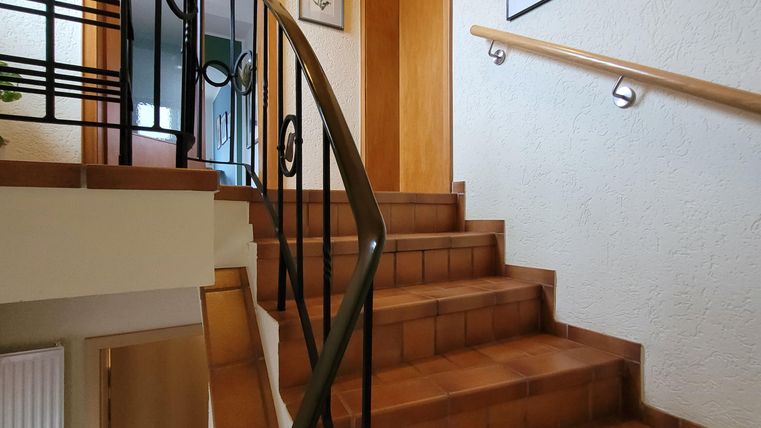 Eine Treppe mit braunen Fliesen und einem handlauf aus Metall. An den Wänden hängen mehrere Bilder und es gibt einen Eingang zu einem Raum oben.