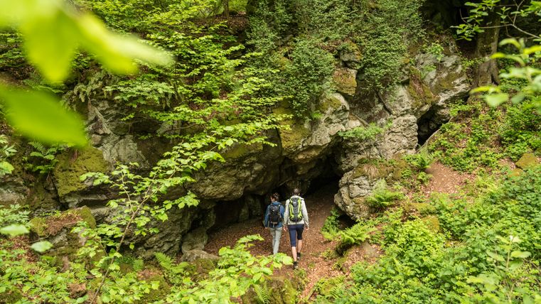 Deux randonneurs entourés de rochers couverts de végétation. Ils se dirigent vers l'entrée d'une grotte.
