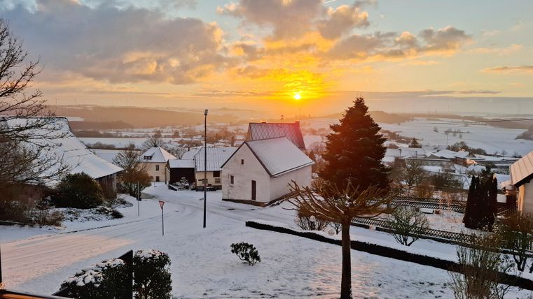 Eine verschneite Landschaft mit Häusern und Bäumen. Im Hintergrund strahlt die Sonne am Horizont.