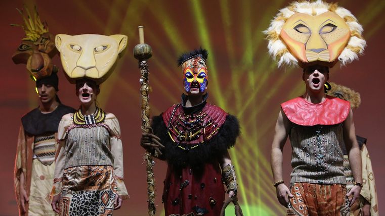 Eine Gruppe von Darstellern in bunten Kostümen mit Tiermasken und einem Stab. Sie stehen auf einer Bühne mit bunten Lichtern im Hintergrund.