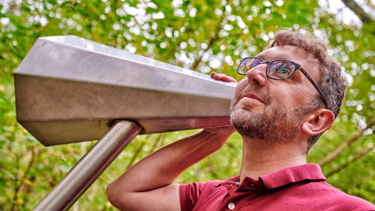 A man listens to a large metal ear trumpet outside.