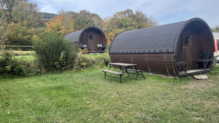 Deux cabanes confortables se trouvent dans une prairie verte, entourées d'arbres. Le temps est nuageux, et il y a un petit coin salon en plein air.