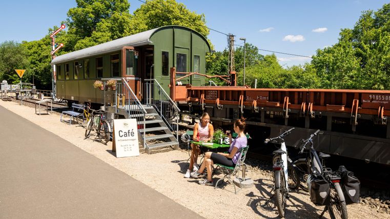 Un café dans un wagon de train aménagé, entouré d'arbres. Deux femmes sont assises dehors et profitent de leur temps.