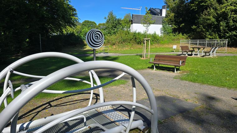 A playground with modern equipment and a lawn in the background. The surroundings are green and it is sunny.