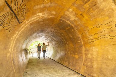 Zwei Personen gehen durch einen tunnelartigen Holzsteg am Wilden Kermeter auf dem Wilden Weg. Die begehbare Konstruktion ist einem hohlen Baumstamm nachempfunden.