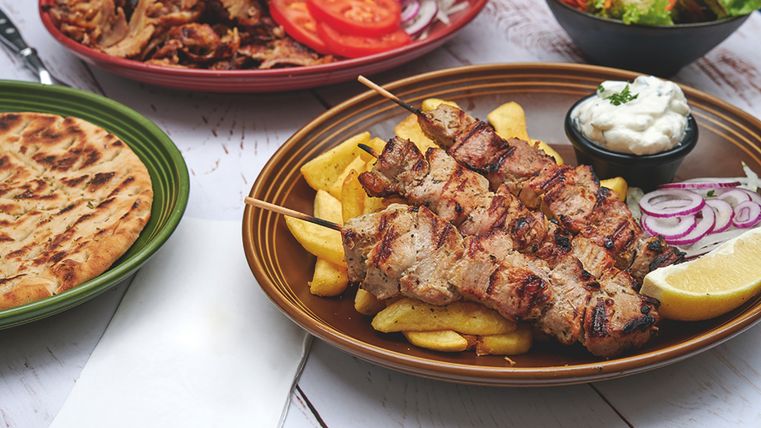 Auf einem Holztisch stehen vier Speisen: Gyros mit Pommes, Fleisch, Brot und ein gemischter Salat