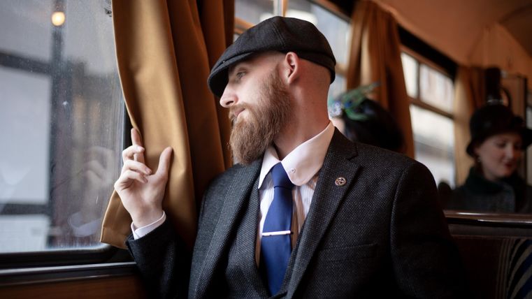 Ein stilvoll gekleideter Mann mit Bart schaut aus dem Fenster eines Zugabteils. Die Atmosphäre ist nostalgisch und elegant.