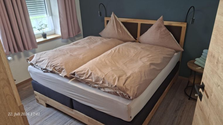 A cozy bedroom with a large double bed and beige bedding. The walls are painted in a dark shade, and there are windows with light curtains.