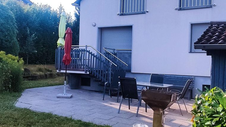 A modern house with a small garden. There are chairs and a grill on the terrace.