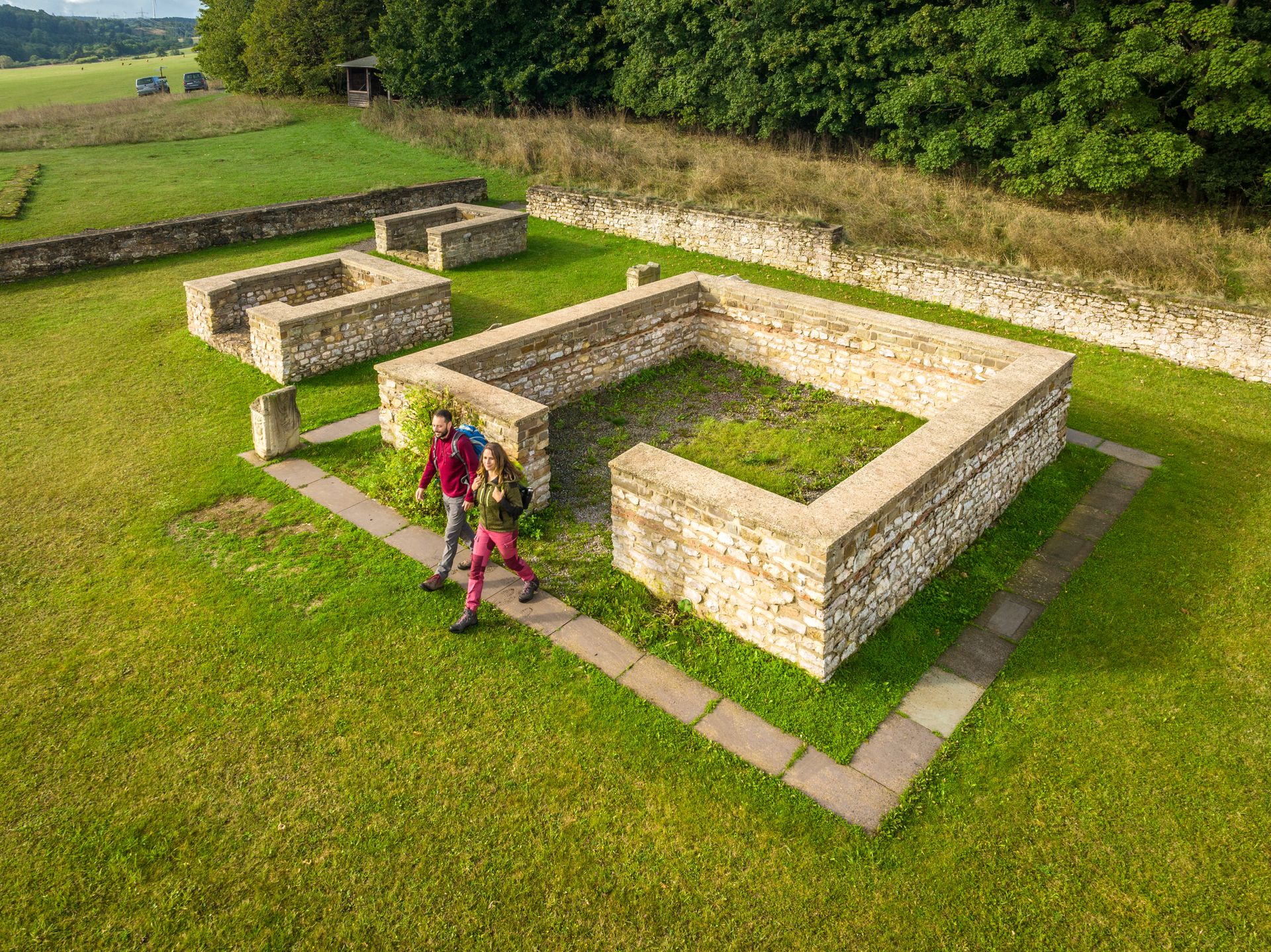 Luftaufnahme des Matronenheiligtums Görresburg bei Nettersheim. Zu sehen sind die freigelegten Grundmauern römischer Kultbauten aus Stein, eingebettet in eine grüne Wiese. Zwei Besucher Wandern vorbei an den zentralen Strukturen. Im Hintergrund verläuft eine Baumreihe entlang einer Mauer.