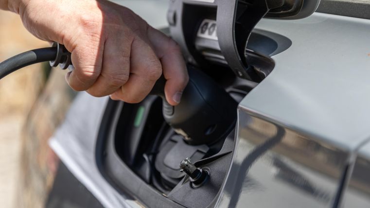 A hand is plugging a charging connector into the charger of an electric car. In the background, blurred natural elements are visible.