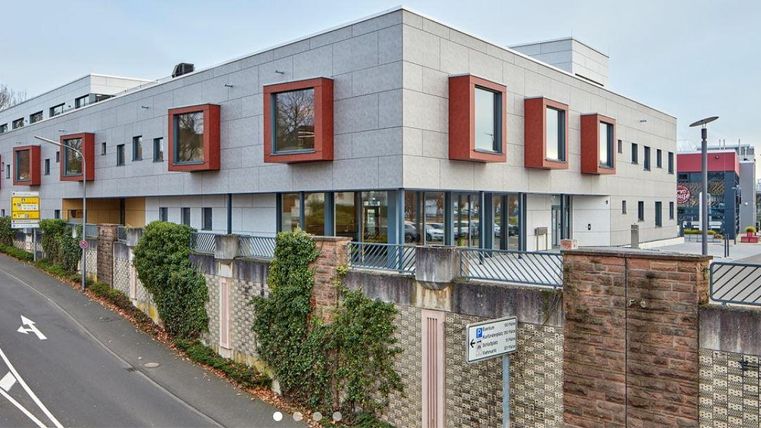 Ein modernes Gebäude mit charakteristischen roten Fenstern. Es befindet sich an einer Straße, umgeben von einer Mauer und grünen Pflanzen.