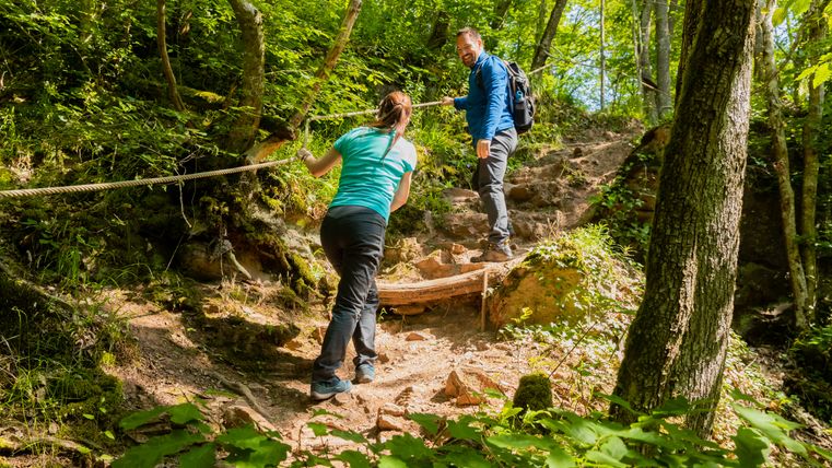 Zwei Personen wandern einen steilen, bewaldeten Pfad hinauf, halten sich an einem Seil fest.