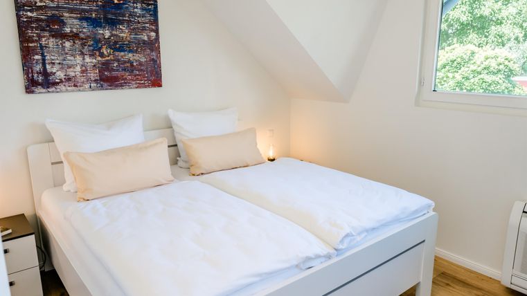 Een lichte slaapkamer met een tweepersoonsbed en witte dekbedden. Aan de muur hangt een modern kunstwerk, en het raam biedt uitzicht op de groene omgeving.