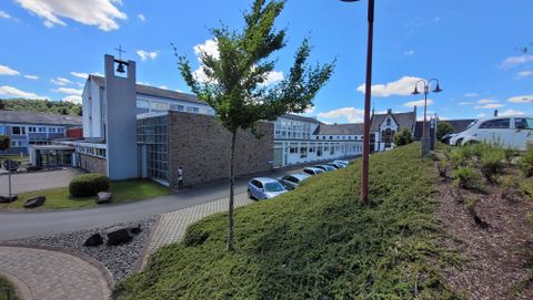 Un bâtiment moderne avec de grandes fenêtres et un environnement soigné. Au premier plan, on voit une colline verte avec des buissons et un ciel bleu clair.