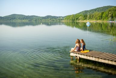 Zwei Kinder sitzen entspannt am Ende eines Holzstegs, der in den ruhigen, klaren Laacher See führt; sie beobachten die kreisförmigen Wellen auf dem Wasser, umgeben von grünen Hügeln und bewaldetem Ufer bei sonnigem Wetter.