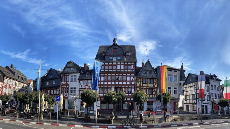 Een mooi historisch stadsplein met verschillende vakwerkhuizen. De lucht is helder en blauw, en er wapperen vlaggen aan de gebouwen.