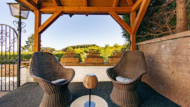 A cozy terrace with two woven chairs and a small table. In the background, a green landscape unfolds under a clear sky.