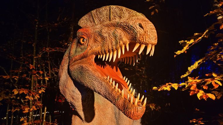 Une tête de dinosaure réaliste se tient parmi des arbres dans l'obscurité. L'environnement est entouré de feuilles d'automne.