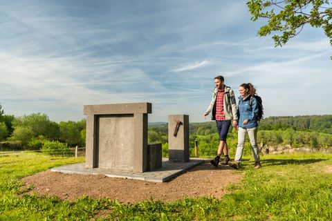 Deux randonneurs passent devant une sculpture en pierre qui ressemble à un trône, sur une colline verdoyante avec un fond boisé.