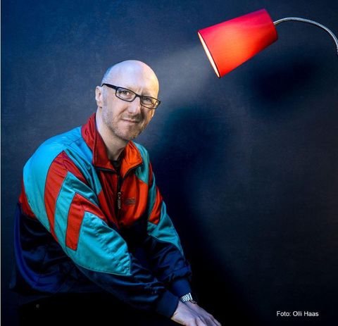 Ein Mann mit Brille sitzt neben einer roten Lampe. Er trägt eine bunte Trainingsjacke und lächelt in die Kamera.