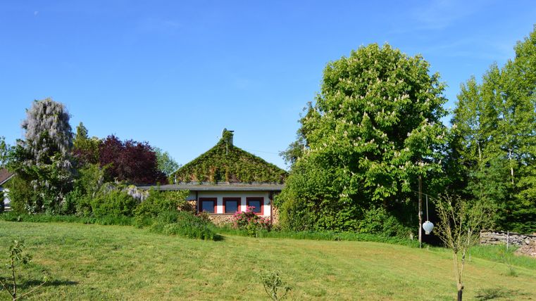 Ein grünes Haus mit einem bewachsenen Dach steht inmitten eines schönen Gartens. Im Hintergrund sind hohe Bäume und ein klarer blauer Himmel zu sehen.