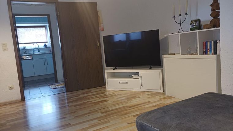 Une salle de séjour confortable avec une télévision et une étagère moderne pour TV. La pièce a un sol en bois clair et mène à une cuisine.