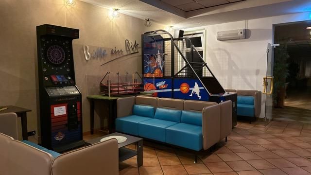 A cozy room with blue seating and a slot machine. In the background, there is a basketball game device and a dartboard.