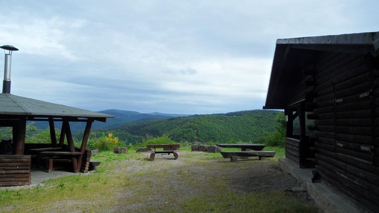 Eine ruhige Landschaft mit einem Holzhaus und einem Unterstand. Im Hintergrund sind grüne Hügel und bewölkter Himmel sichtbar.