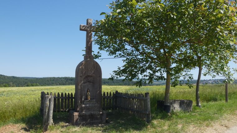 A stone wayside cross stands next to a tree on a country lane, surrounded by a wooden fence. Fields and a forest can be seen in the background.