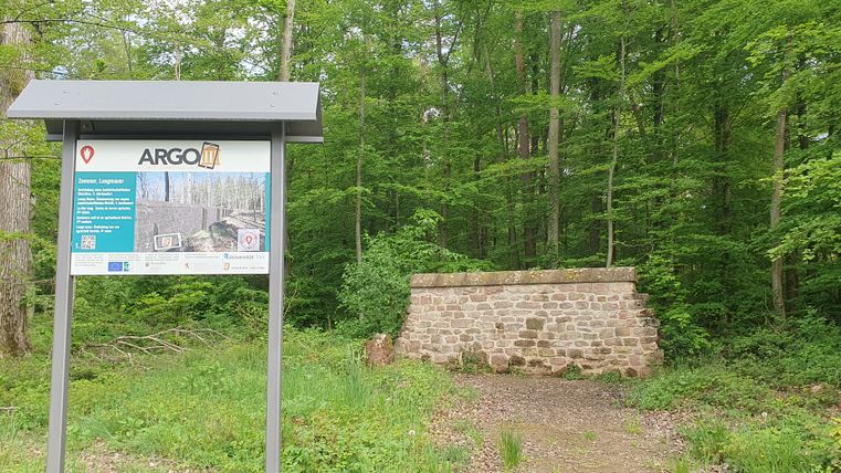 Eine Informationstafel vor einer Steinmauer inmitten eines Waldes