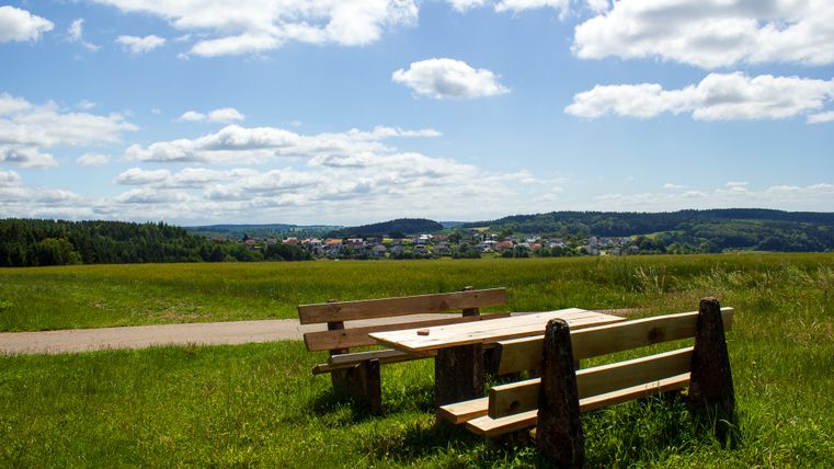 Houten tafel en banken op een weiland met uitzicht op Neidenbach en beboste heuvels op de achtergrond.