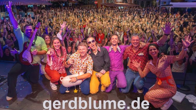 Eine fröhliche Gruppe von Menschen steht auf einer Bühne und posiert für das Foto. Im Hintergrund ist eine große Menge von Zuschauern zu sehen, die begeistert applauden.