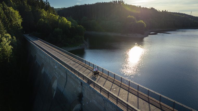 In the foreground, two cyclists are riding on a dam in sunny weather. In the background, the Oleftalsperre, on which the sun is reflected, is surrounded by a forest. 