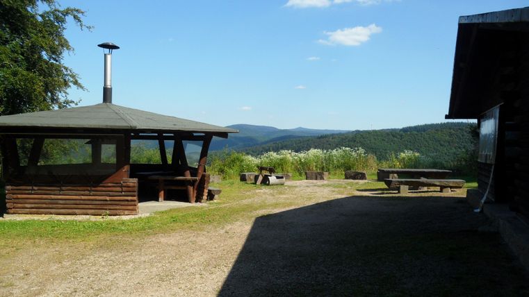 Une vue spacieuse sur un paysage vallonné avec un coin barbecue et un kiosque en bois. Le ciel est clair et l'environnement est verdoyant.