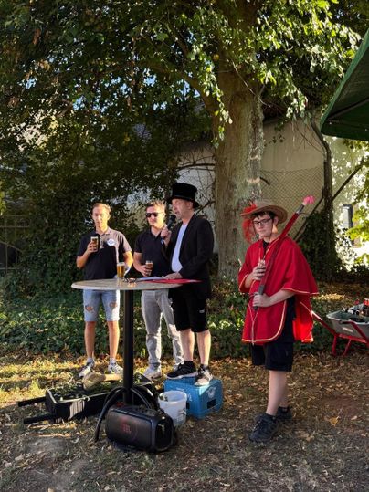 Eine kleine Veranstaltung im Freien mit mehreren Personen. Ein Mann trägt einen Zylinder und spricht zu einer Gruppe, während ein kindlicher Teilnehmer eine rote Kostümjacke und einen Stock hält.