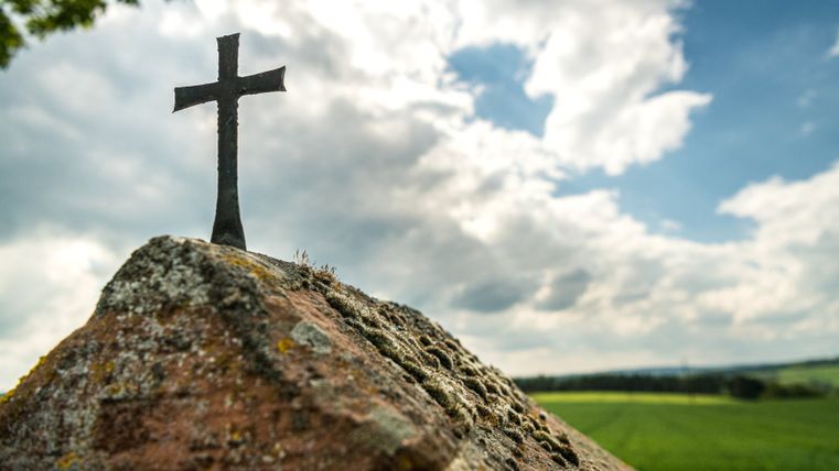 Een klein, zwart kruis op een met mos bedekte steen voor een bewolkte hemel.