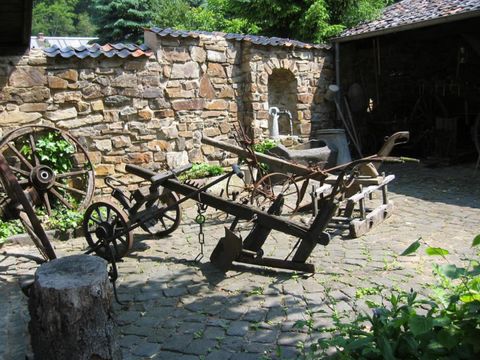 Een historische boerderij met oude landbouwmachines in een geplaveide binnenplaats. Op de achtergrond ziet men een stenen muur en enkele bomen.