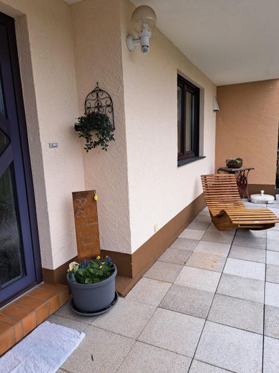 Een uitnodigende entree met een houten ligstoel en een bloembak. De muren zijn licht gehouden en er is een decoratieve wanddecoratie.