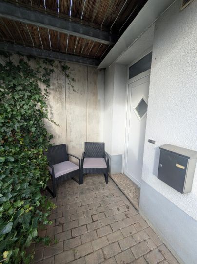 Twee stoelen staan voor een voordeur, omgeven door een muur met klimop. Naast de deur staat een brievenbus.