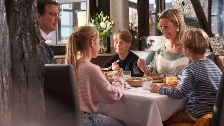 Een gezin zit samen aan tafel en geniet van een maaltijd. Ze lachen en hebben een fijne tijd met elkaar.