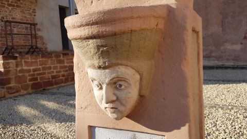 Een kunstzinnige sculptuur met een gezicht en een hoed van steen. De figuur staat tegen een muur in een historisch rijke omgeving.
