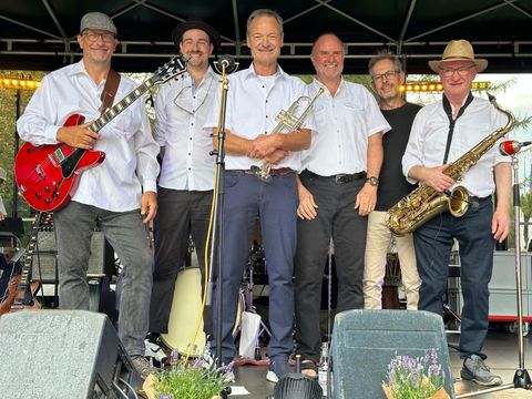 Een band staat op een podium en poseert voor een foto. Ze dragen witte shirts en houden muziekinstrumenten vast.