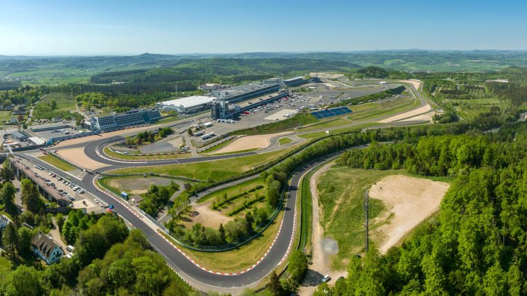 Eine Luftaufnahme einer Rennstrecke umgeben von grünen Hügeln. Im Hintergrund sind Gebäude und Parkplätze sichtbar.
