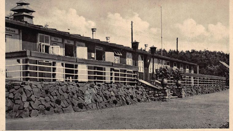 Un bâtiment historique avec plusieurs fenêtres et une véranda. Entouré d'un terrain soigné et d'arbres en arrière-plan.