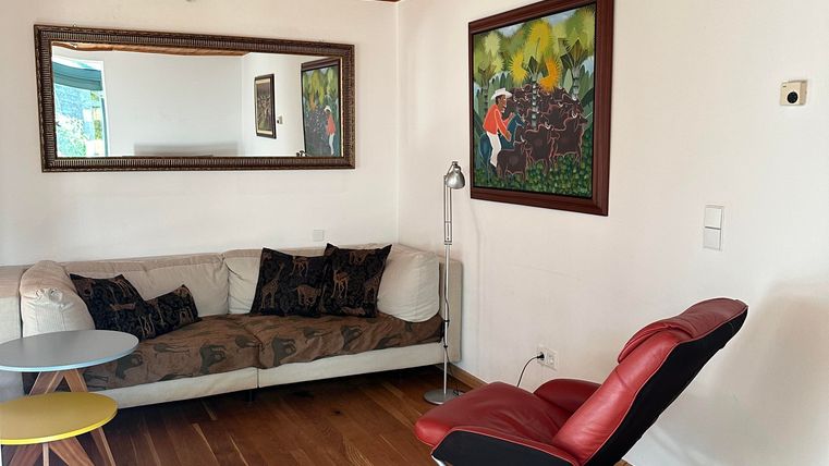 A bright room with wooden floors and a sofa. Paintings hang on the wall, and a comfortable chair stands in the room.