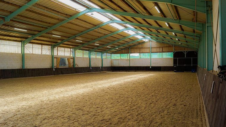 Une grande carrière de dressage bien éclairée avec un sol sablonneux. Les murs sont en bois et le plafond est en structure d'acier.