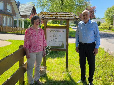 A couple stands next to a sign that reads "Holiday Home" in an idyllic street. In the background, green trees and typical North German houses can be seen.