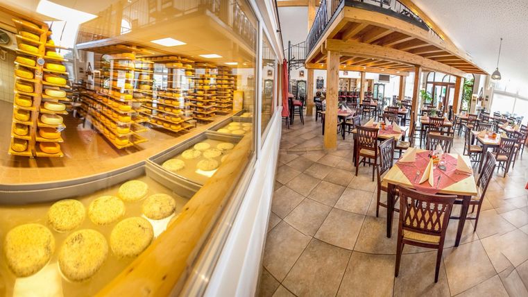 Een uitnodigend restaurant met houten decor en uitzicht op rijpe kaas. De tafels zijn voorbereid voor gasten en er zijn planken vol kaas op de achtergrond.
