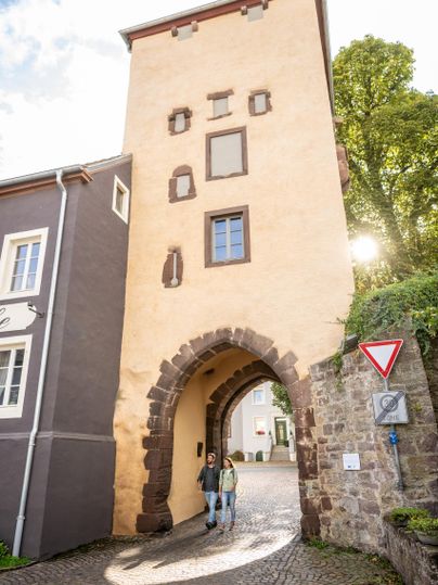 Een historisch poort van steen met een boogvormige ingang. Twee personen lopen daaronder door op een geplaveide weg.
